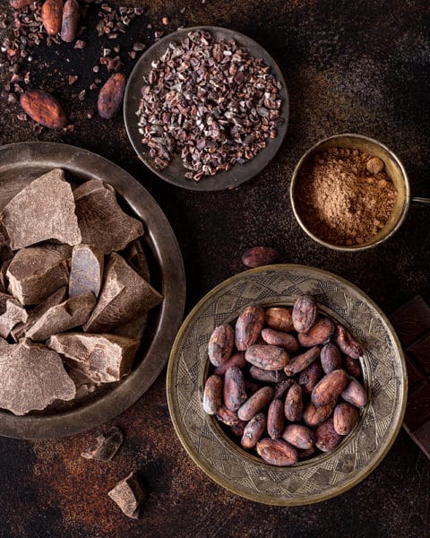 Ein Tisch mit Kakaobohnen in einer Schüssel, Kakaopulver in einer kleineren Schüssel. Kakaonibs auf einem kleinen schwarzen Telller und Schokoladenbrocken in einer grossen Schüssel. Zur Verzierung liegen Kakaobohnen, Pulver, Nibs und Schokolade auf dem TIsch.