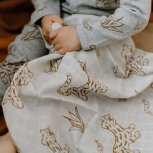 Kleiner Junge im grauen Body mit Senfgelben Leoparden hält ein weisses Nuschi ebenfalls mit senfgelben Leoparden und Pflanzen in der Hand