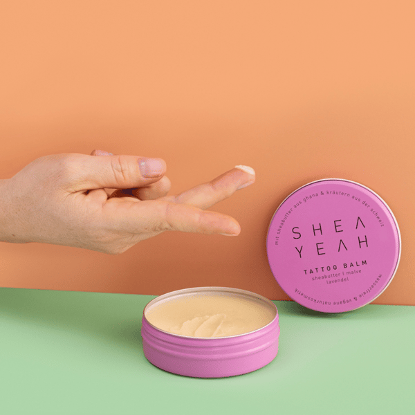 Finger hält etwas Tattoo Balm über der offenen Verpackung und vor farbigem Hintergrund.