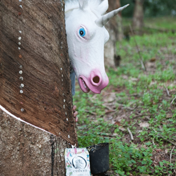 Ein Mann mit einer Einhorn-Maske versteckt sich im Wald hinter einem Baum. Vor dem Baum steht eine Tüte einhorn Kondome.