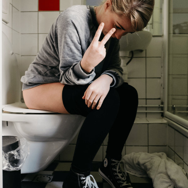 Blonde Frau sitz auf dem WC und zeigt der Kamera das umgekehrte Peace Zeichen. Sie trägt einen grauen Pulli, eine schwarze Leggins und schwarze Converse.